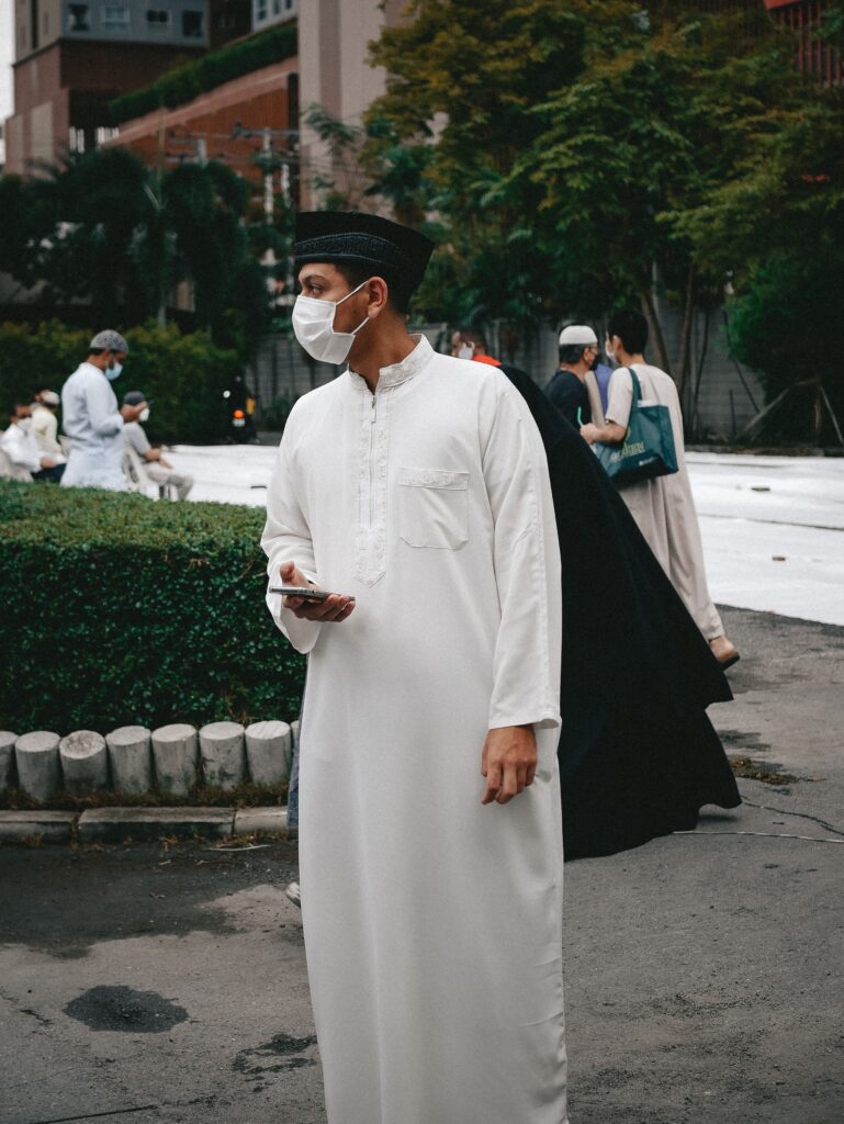 Asian man wearing a face mask and traditional attire holding a smartphone outdoors.